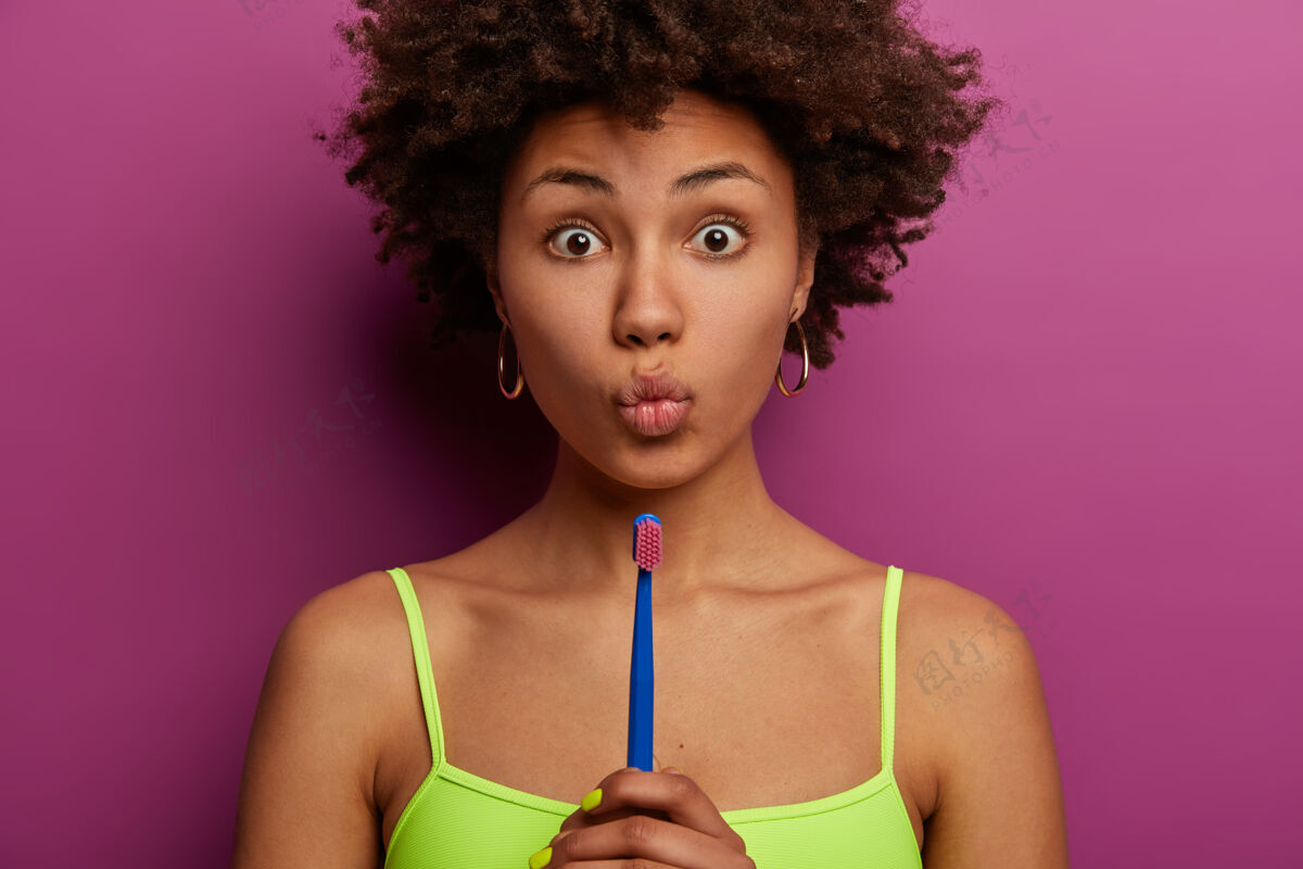 非洲自然美的惊讶的少数民族妇女的剪短的形象 保持嘴唇折叠 拿牙刷 去清洁牙齿 穿衬衫 模型在充满活力的紫色墙壁口腔卫生概念惊喜自然肩膀