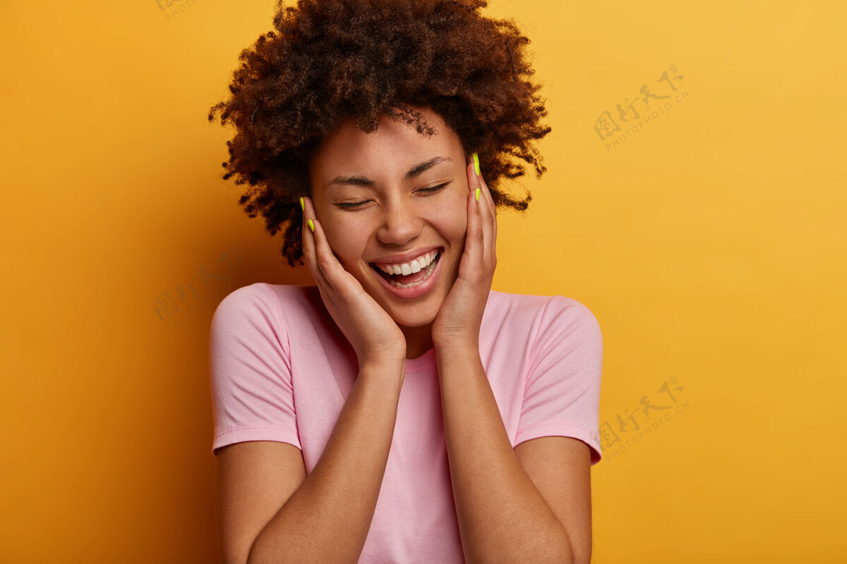 美甲快乐的卷发女人的特写肖像触碰脸颊 有真诚积极的微笑 闭上眼睛和被逗乐 笑从一些搞笑的东西 穿着随意 在黄色的墙壁上微笑休闲牙齿
