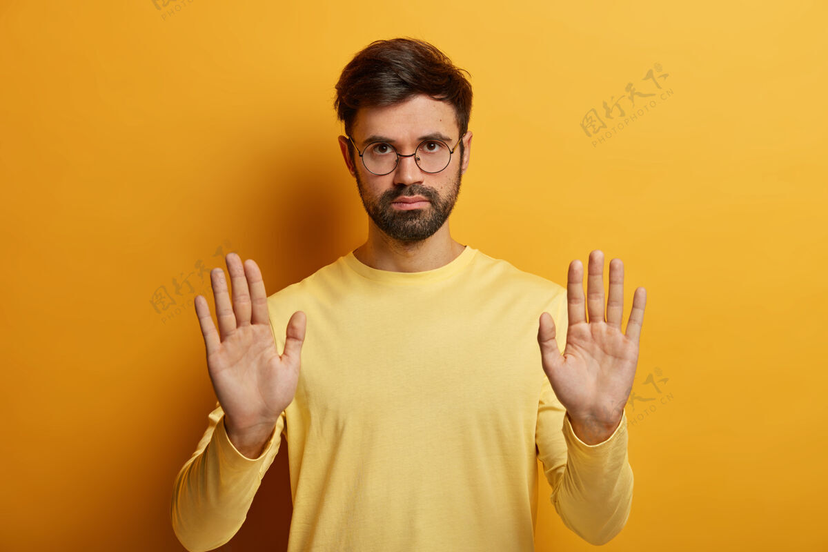 严肃严肃的未刮胡子男子肖像作停车标志 表示限制 拒绝或拒绝 要求不要打扰他戴圆眼镜和隔离在黄色墙上的套头衫有警告的表情拒绝禁止白种人