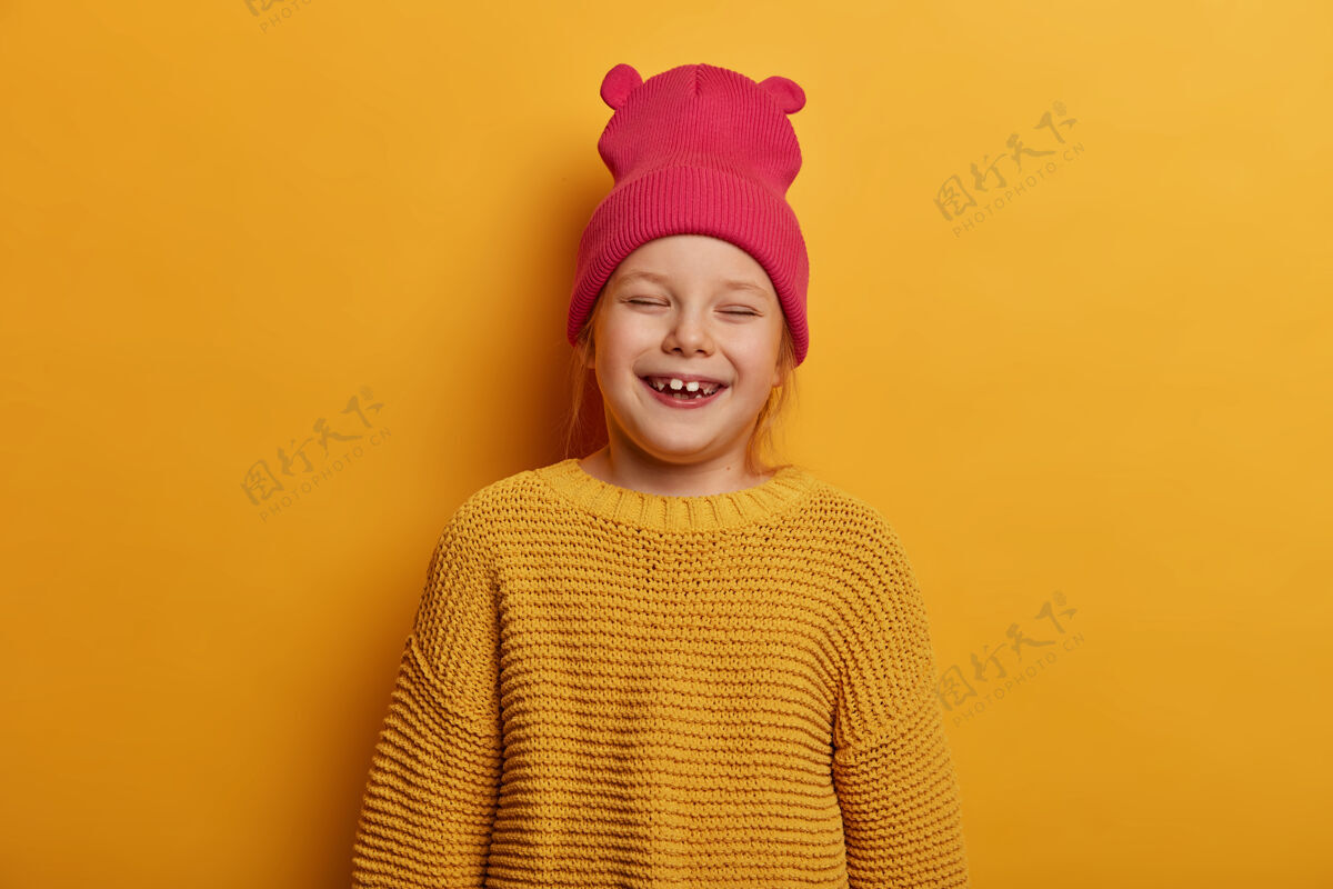 快乐可爱的小女孩笑得很开心 很高兴收到父母的新玩具娃娃 闭上眼睛 在室内玩得很开心 戴着带耳朵的帽子和宽松的针织毛衣 隔着黄色的墙快乐乐观编织