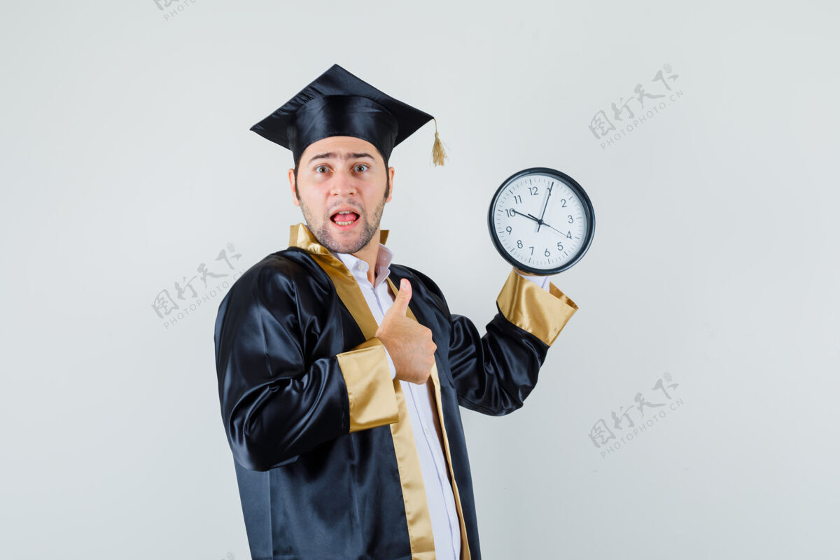 帅哥拿着挂钟的年轻人 在毕业生制服的前视图中竖起大拇指肖像钟墙