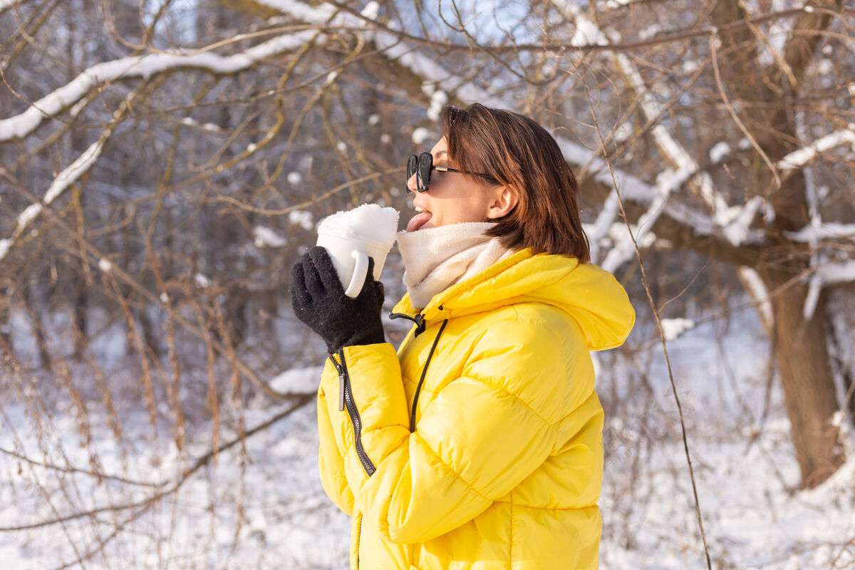 森林美丽的年轻开朗的女子在雪景的冬日森林里戴着墨镜拿着装满雪的杯子玩得开心冬天美丽肖像