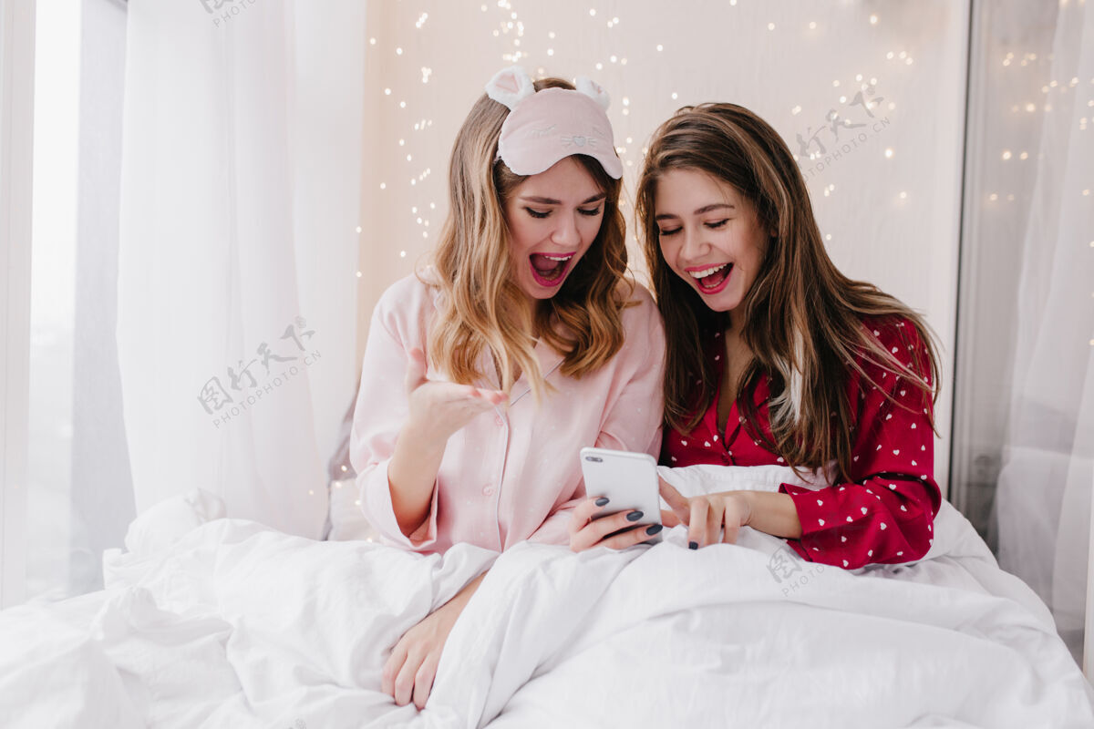 室内穿着时尚睡衣的漂亮女孩在发短信迷人的年轻女士一起坐在床上看智能手机屏幕的室内写真年轻快乐电话