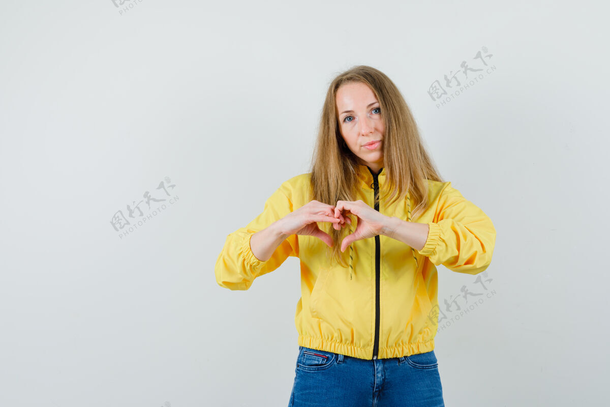 成人身穿黄色棒球服和蓝色牛仔裤的年轻女子 展现出心形 看起来迷人 正面视图漂亮可爱心