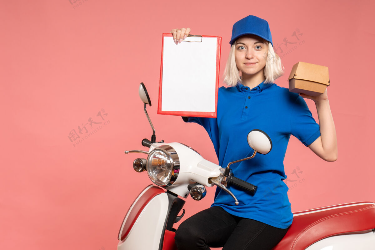 工作前视图骑着自行车的女快递员 粉红色的小食品包送货食品工人