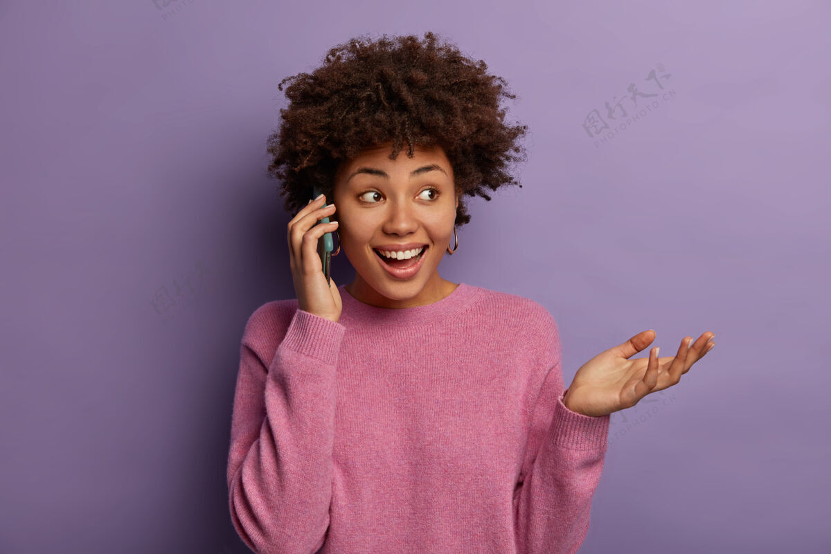 外表快乐的民族妇女在电话里交谈 目光移开 做手势 用印象讨论她去剧院的事 穿着休闲的套头衫 聚精会神地站在一边 在紫色的墙上有着孤立的牙齿般的微笑欢乐女性微笑