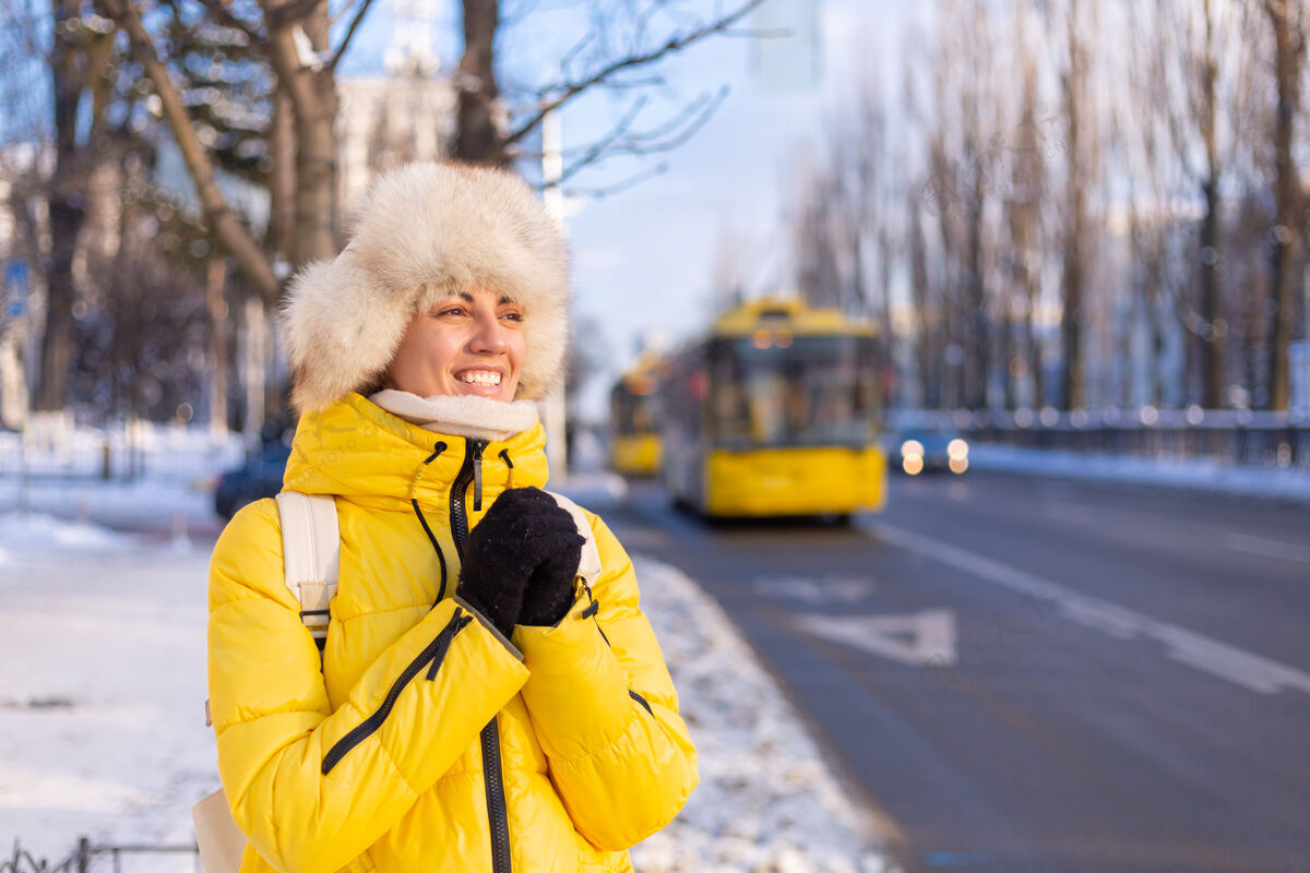 女孩一个穿着暖黄色夹克和西伯利亚俄罗斯帽子的快乐女人在雪城街道上等待公共汽车的冬季肖像乐趣城市女性