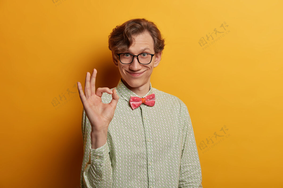 模特笑容可掬的男极客用手指做手势 说工作不错 对建议很满意 戴着眼镜 穿着正式的衬衫 看起来很高兴 孤立在黄色的墙上积极人脸
