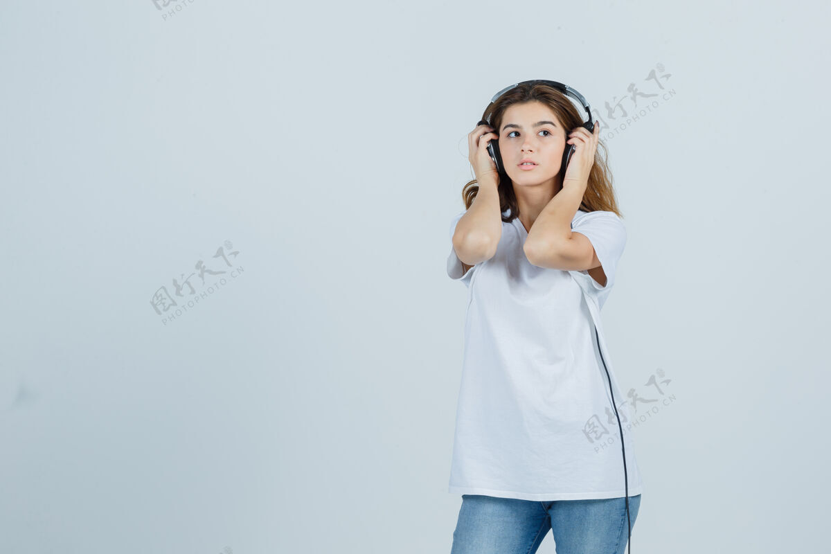 乐趣年轻的女性手放在她的耳机上 穿着白色t恤 牛仔裤 看起来很梦幻 正面视图女人漂亮女性