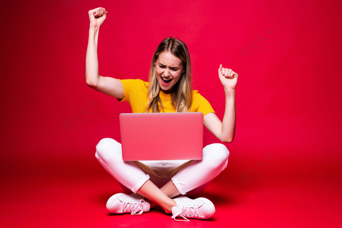 女士快乐的年轻女子 抱着胜利的姿态 盘腿坐在地板上 在红墙上用笔记本电脑成功电脑坐着