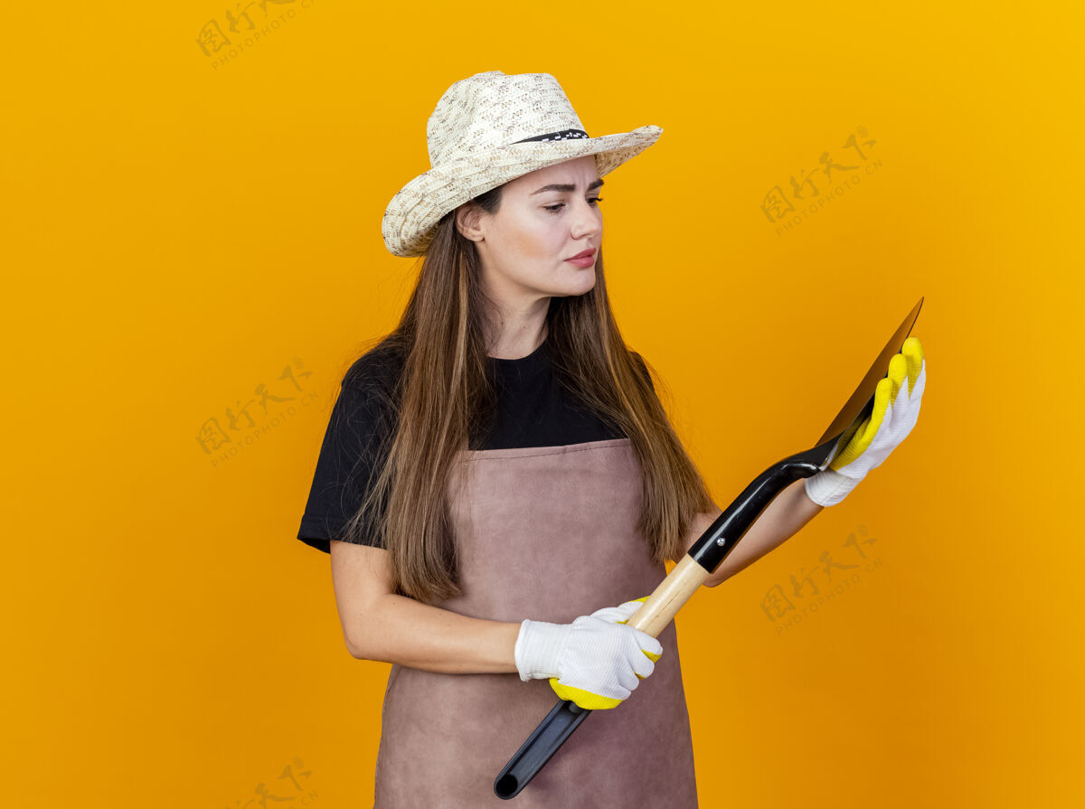花园想着美丽的园丁女孩穿着制服 戴着园艺帽 戴着手套 拿着黑桃看着孤立的橙色背景女孩帽子黑桃
