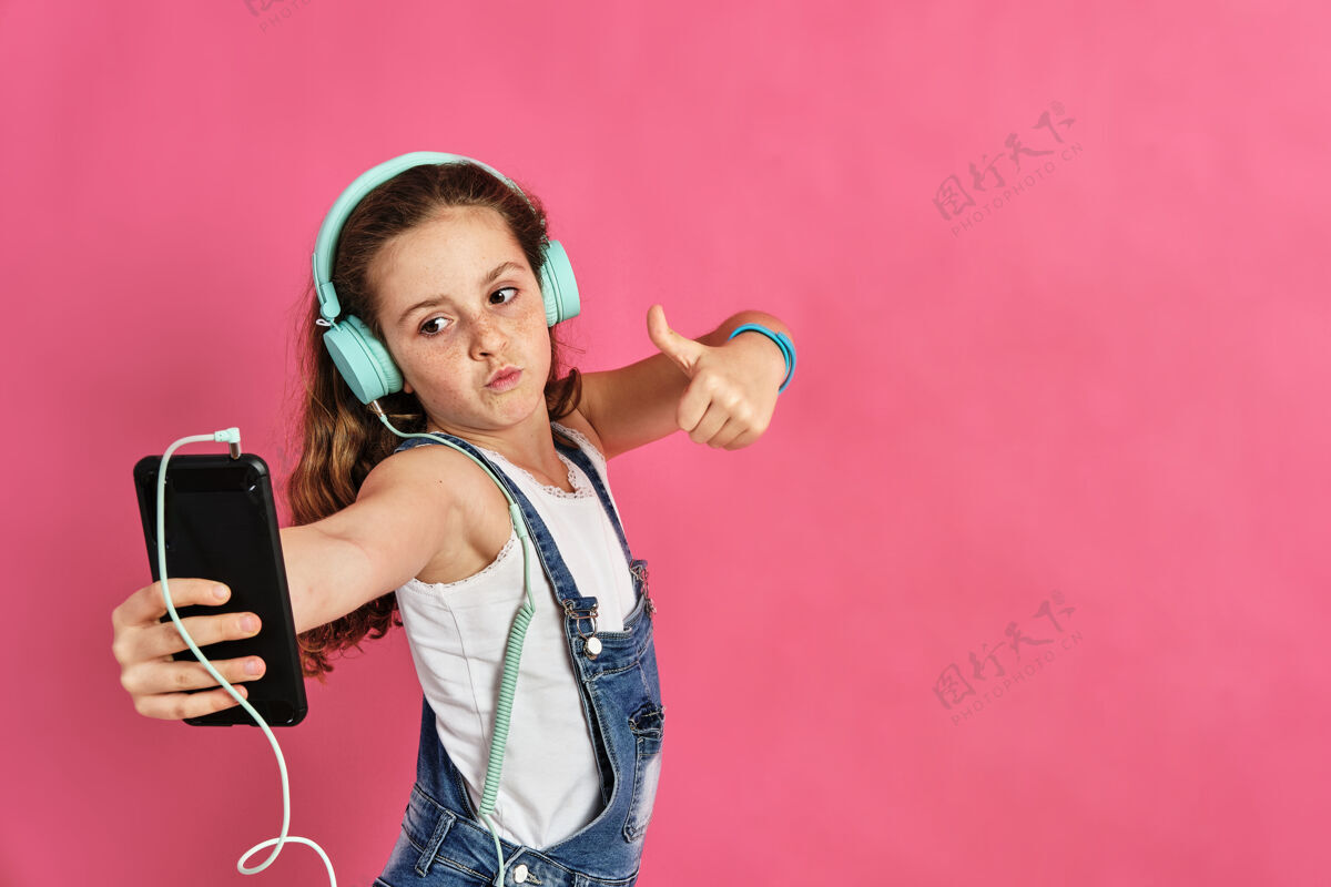 黑发可爱的小女孩在粉红色的墙上用手机和耳机听音乐聪明音乐年轻