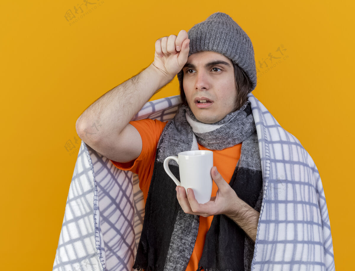 帽子看着身边虚弱的年轻病夫戴着冬天的帽子 披着方格围巾 手里拿着一杯茶 手放在额上 隔离在橙色的背景上额头壁板拿着