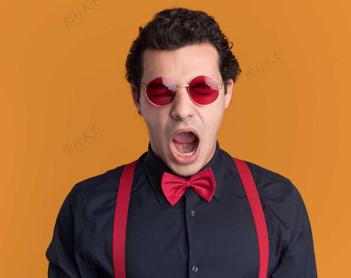领带愤怒的时髦男人戴着蝴蝶结 戴着眼镜和吊带 站在橙色的墙上大喊大叫眼镜蝴蝶结时髦