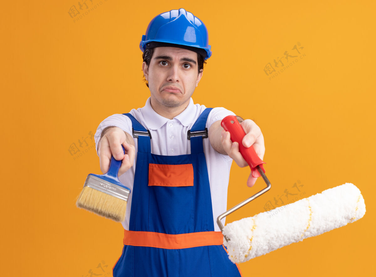 手持年轻的建筑工人身穿建筑制服 戴着安全帽 拿着油漆辊和刷子 站在橙色的墙上 带着悲伤的表情看着前方滚轴安全悲伤
