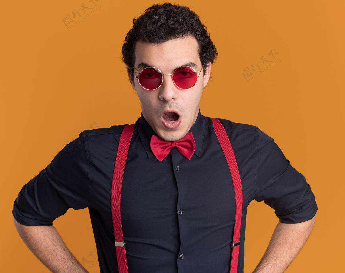 领带愤怒的时髦男人戴着蝴蝶结 戴着眼镜和吊带 站在橙色的墙上 看着前面不高兴男人吊带眼镜
