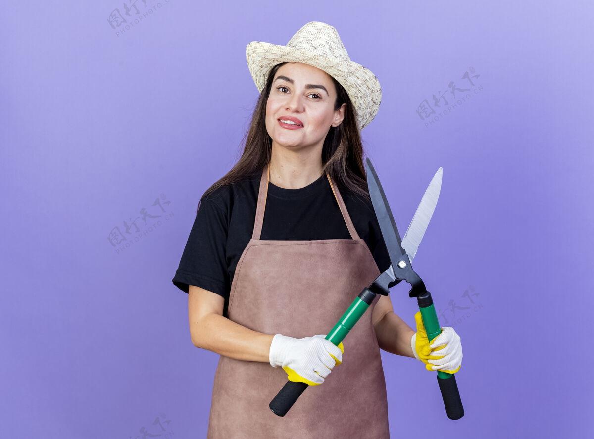 园艺微笑美丽的园丁女孩穿着制服 戴着园艺帽和手套 拿着剪子孤立在蓝色背景上制服手握女孩