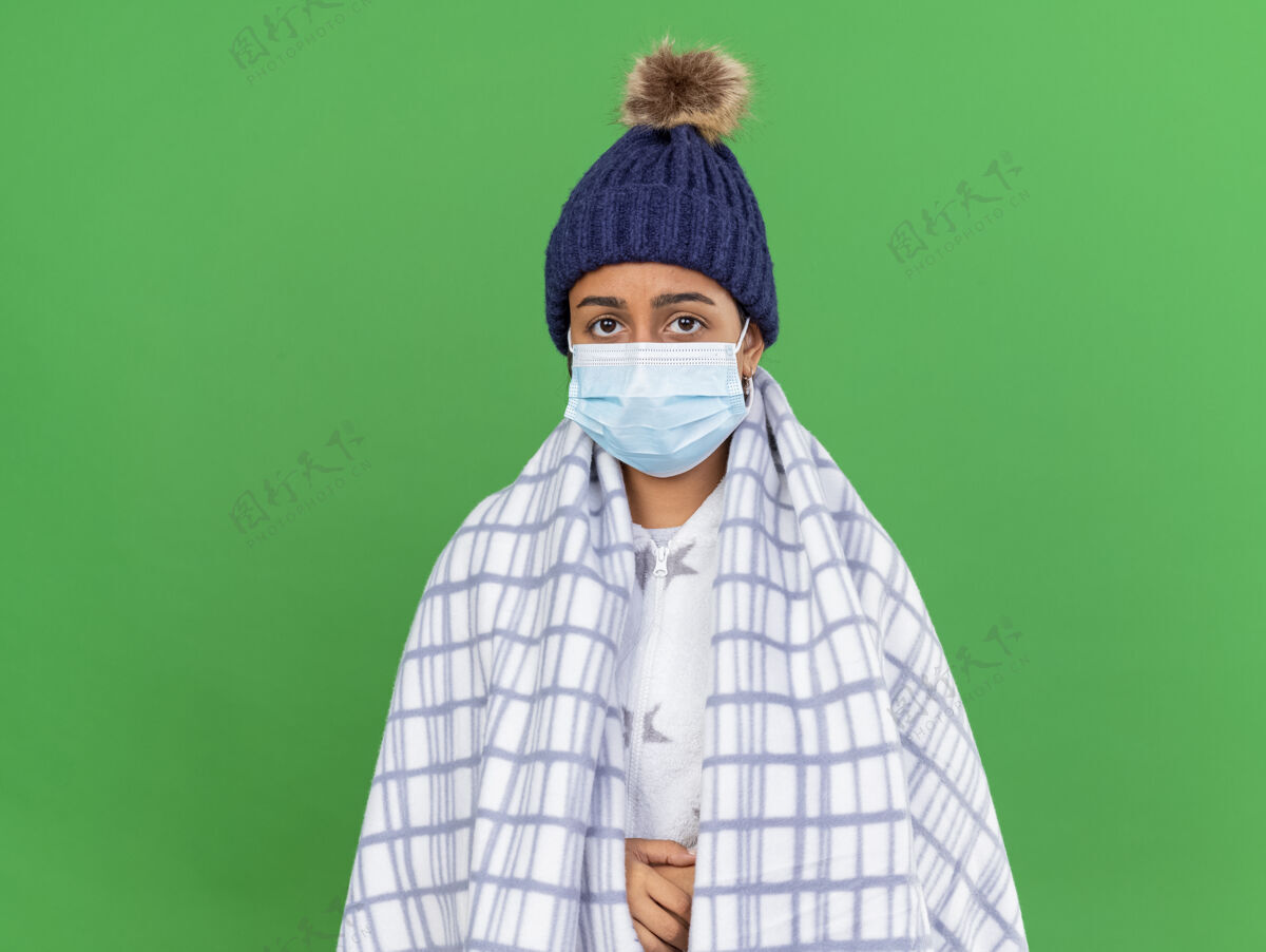 年轻年轻的生病女孩戴着冬天的帽子 戴着围巾和医用口罩 在绿色背景上用格子布包裹着面具格子围巾