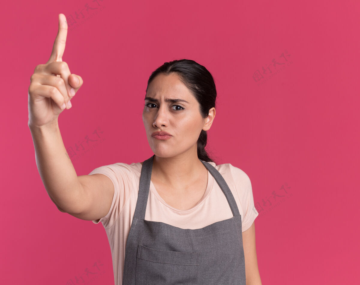 目录年轻漂亮的女理发师围着围裙看着前面 严肃的脸上露出食指警告站在粉红色的墙上围裙年轻手指