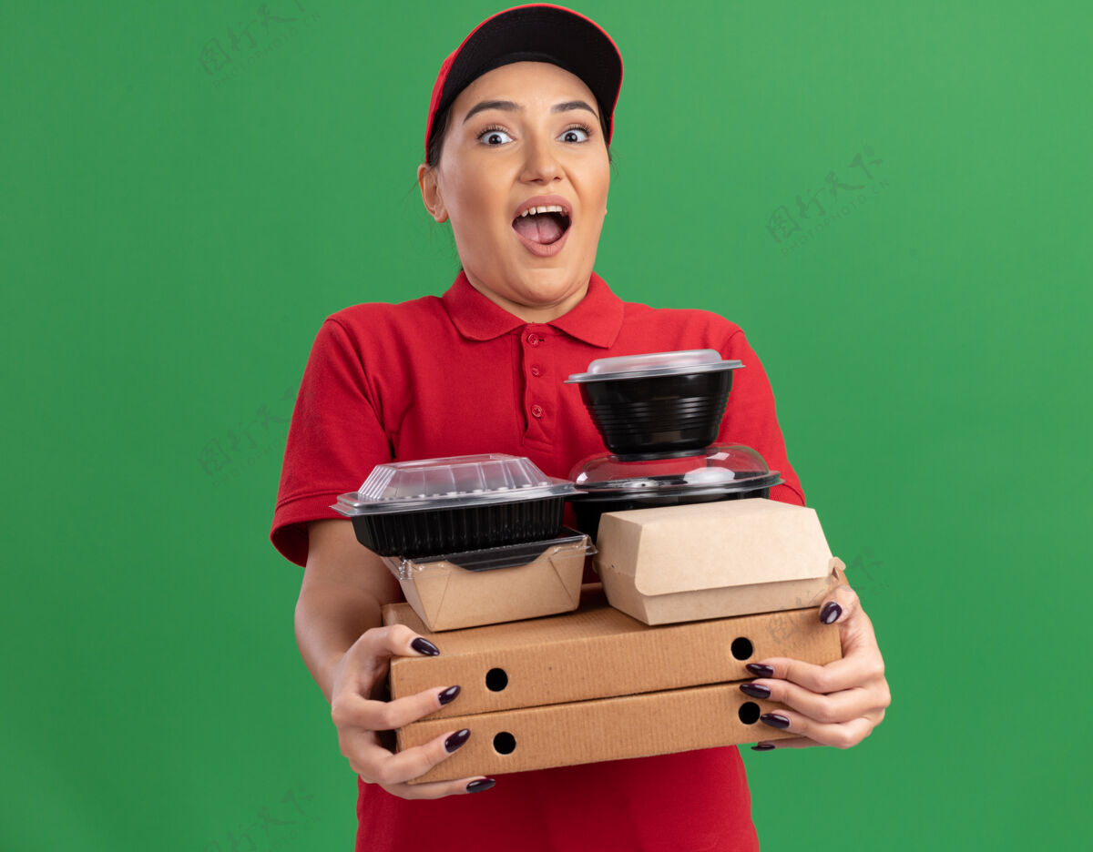 制服年轻的送货员身穿红色制服 头戴鸭舌帽 手里拿着披萨盒和食品包 站在绿色的墙边惊讶地看着前面拿着帽子送货