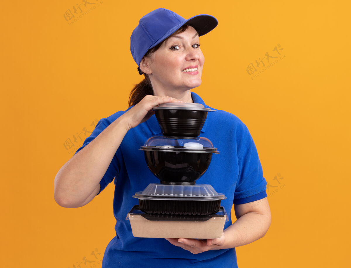 年龄身穿蓝色制服 头戴鸭舌帽的中年女送货员站在橙色的墙上 手里拿着一叠食品包 面带微笑地看着前面盒子站着微笑