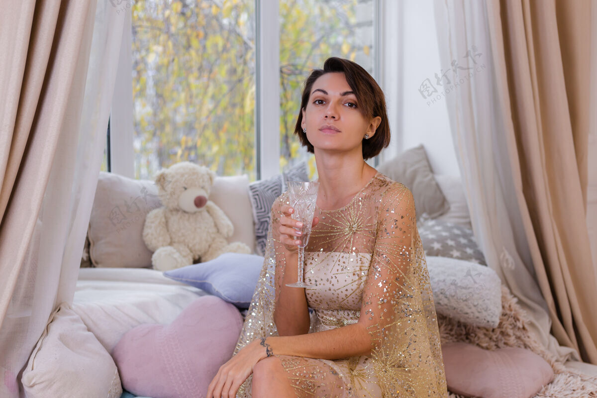 枕头女人在家舒适的房间里穿着圣诞闪亮的晚礼服 捧着一杯香槟头发短裤窗户