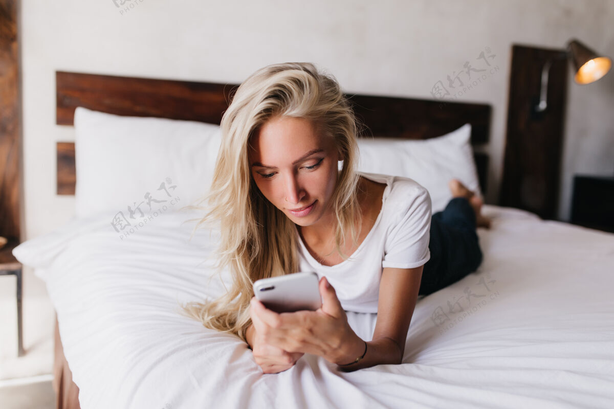 欢乐感兴趣的晒黑女人躺在床上 看着手机屏幕令人惊叹的金发女人在她舒适的卧室冷室内镜头公寓睡眠和平