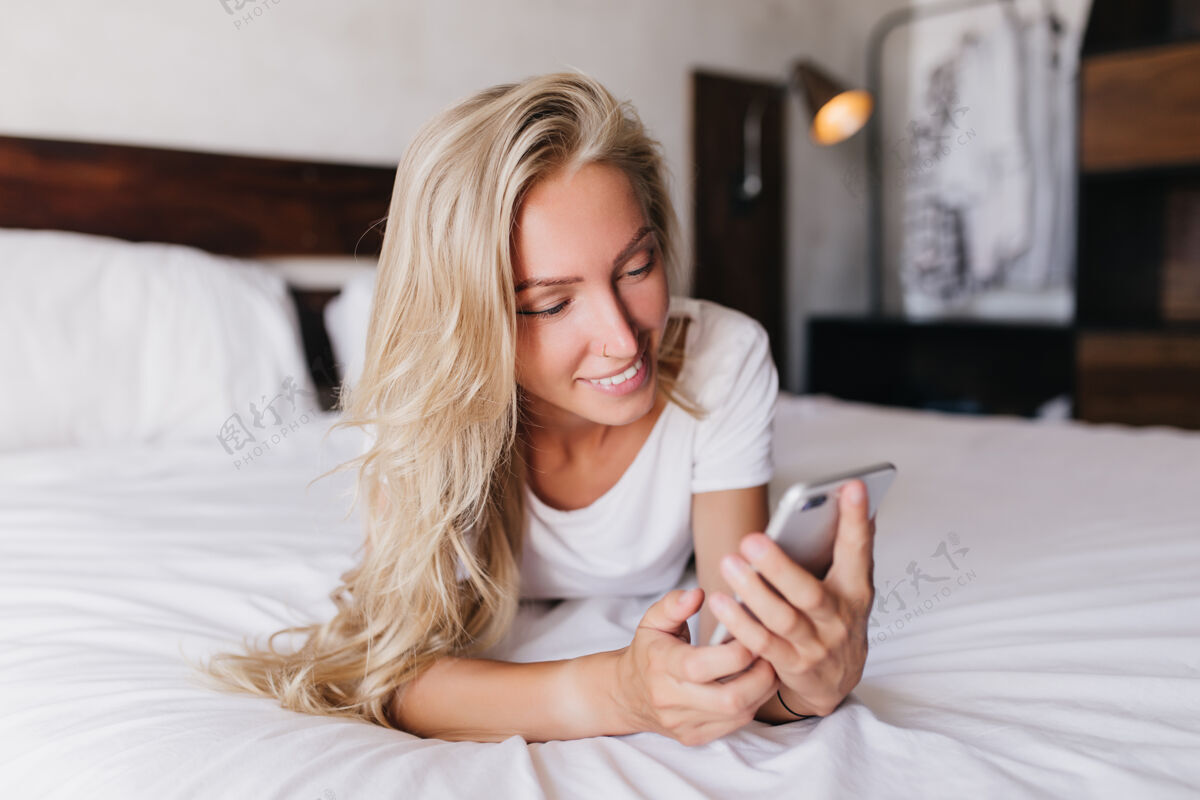 公寓微笑迷人的女人躺在床上拿着电话漂亮的金发女人在卧室里发抖 发短信灯累了手机