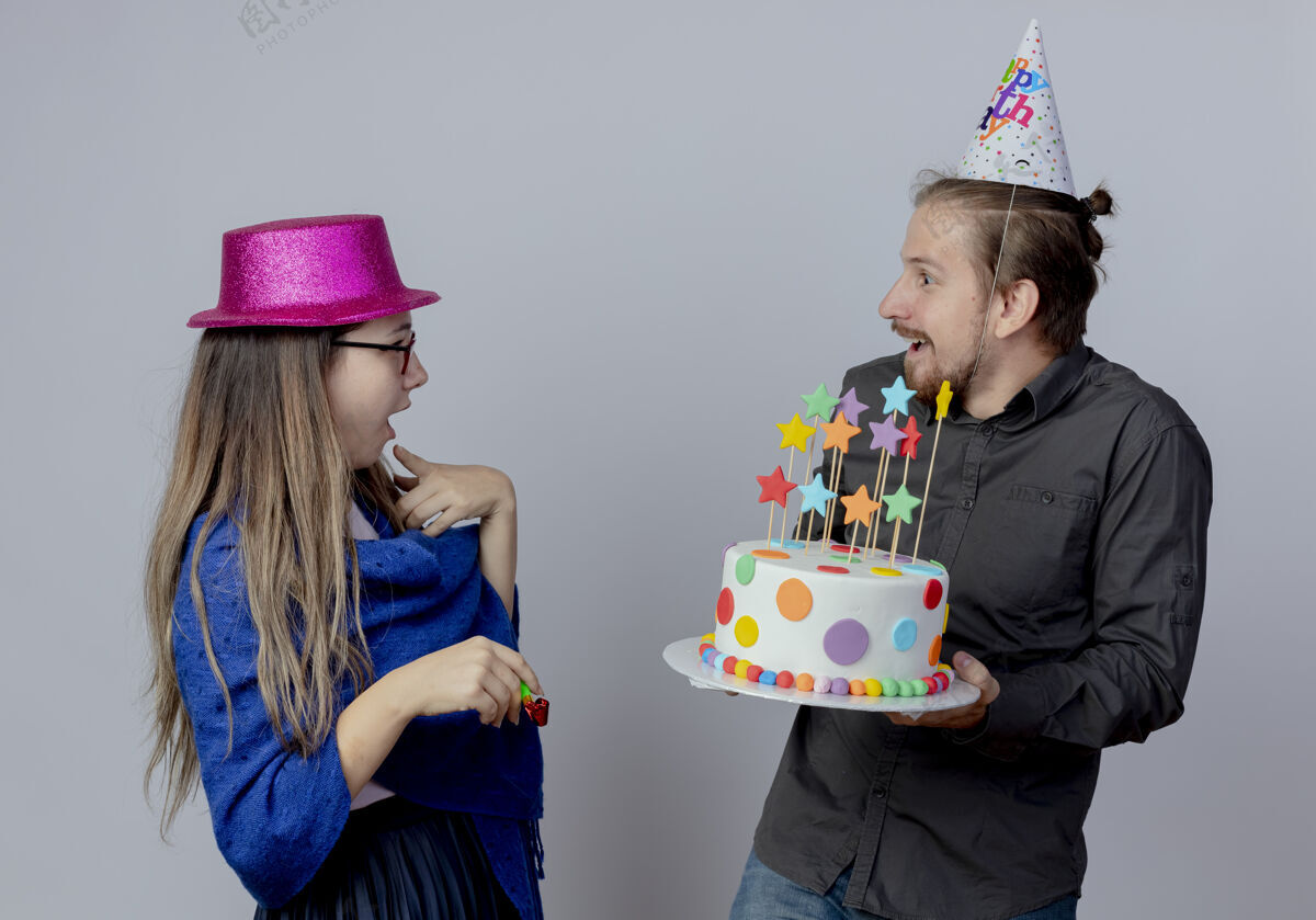 蛋糕惊讶的年轻夫妇看着对方戴着粉红色帽子的戴眼镜的女孩拿着口哨 戴着生日帽的帅哥拿着蛋糕隔离在白色的墙上穿手持眼镜