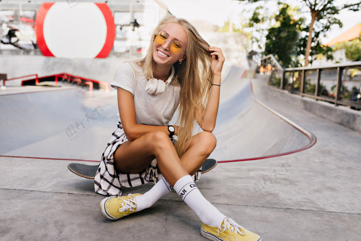 城市可爱的溜冰女玩着她的金发穿着黄色鞋子坐在滑板上的漂亮女模特的户外肖像背景微笑活跃
