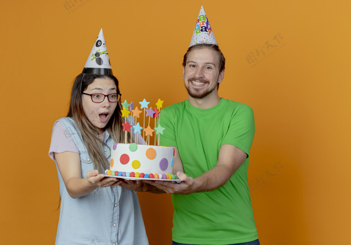 帽子微笑着戴着派对帽的帅哥拿着生日蛋糕 惊讶的年轻女孩戴着派对帽拿着并看着蛋糕年轻蛋糕抱
