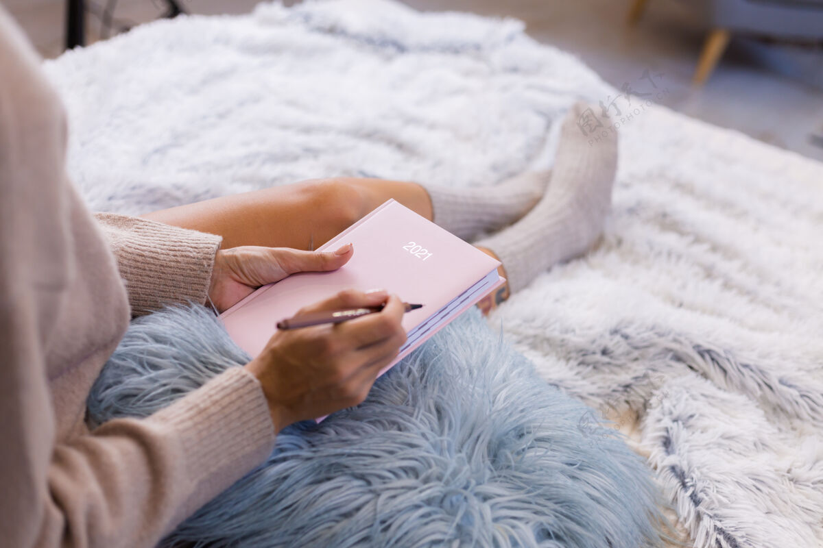 钢笔穿着羊毛袜子和毛衣的女人 粉色笔记本上写着2021 臀部有大纹身女人坐在卧室的家里的床上家庭作业毯子早晨