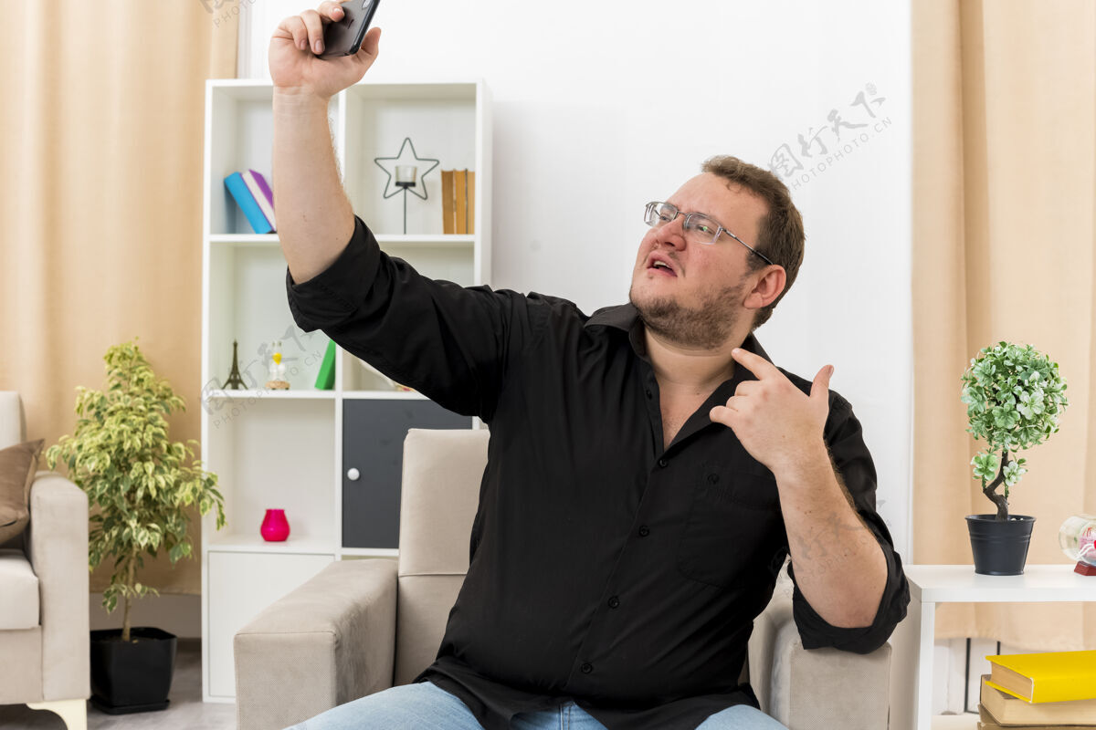 信心自信的成年斯拉夫男子戴着眼镜坐在扶手椅上指着自己 看着客厅里的手机自拍里面自拍采取