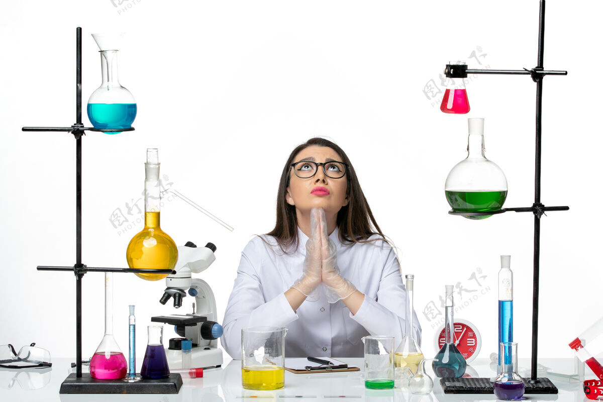 容器前视图穿着白色医疗服的女化学家坐在不同的解决方案上祈祷白色背景科学病毒冠状病毒大流行实验室烧杯溶液实验室