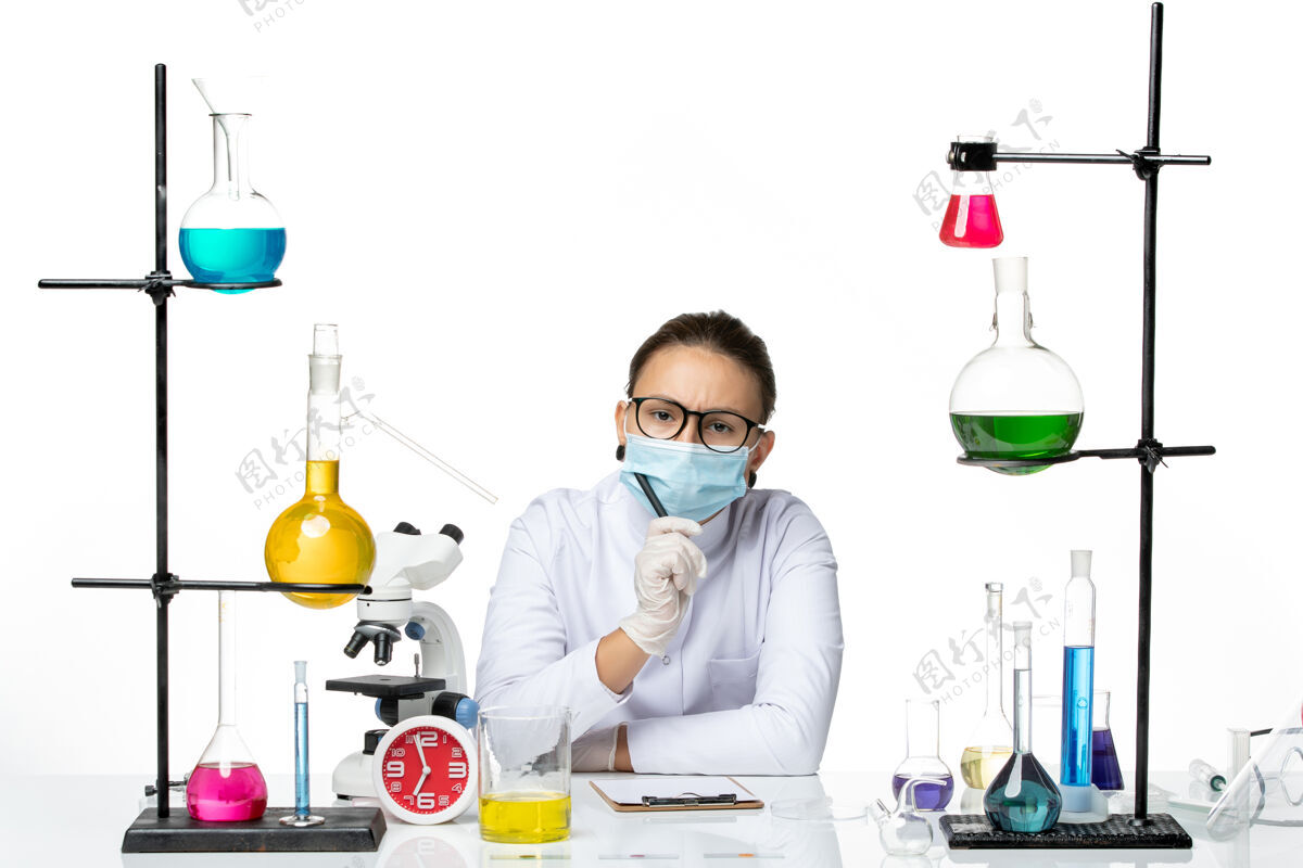 前面前视图穿着医疗服的女化学家 戴着面罩 坐着 拿着溶液 在浅白色背景上写笔记 溅起实验室病毒化学冠状病毒视图解决方案实验室