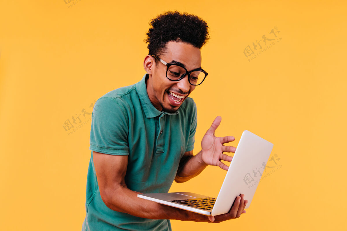 休闲惊讶的卷发男学生看着笔记本电脑屏幕非洲自由职业者戴眼镜的室内照片积极肖像男孩