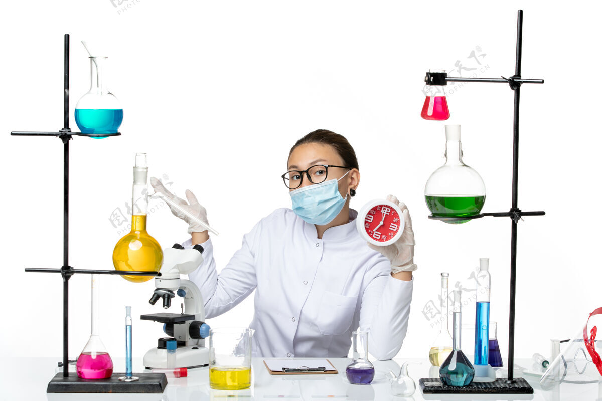 医学前视图：身穿医疗服的女化学家 戴着面罩 手持浅白色背景上的红色时钟病毒实验室化学冠状病毒-飞溅喷溅烧杯医生
