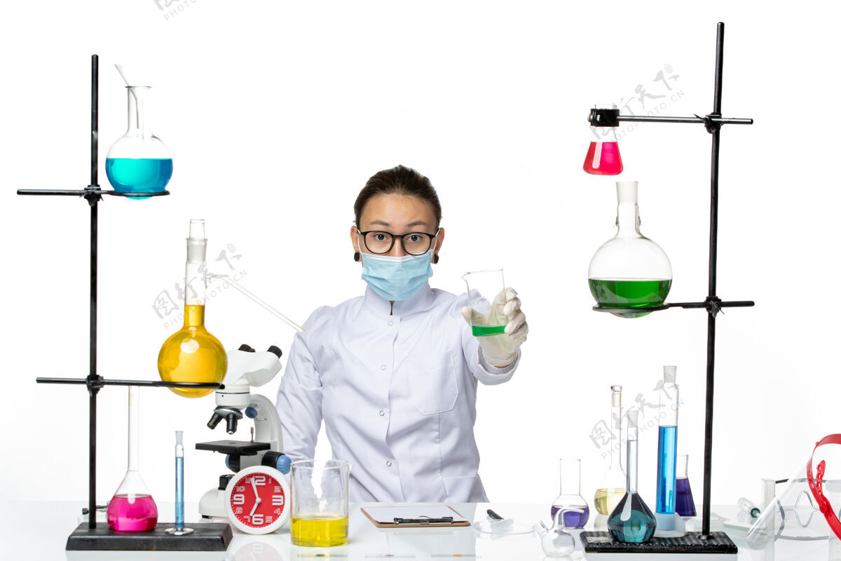 烧杯前视图穿着医疗服的女化学家 戴着面罩 在浅白色背景上拿着绿色溶液 溅起实验室病毒化学covid-病毒保持化学