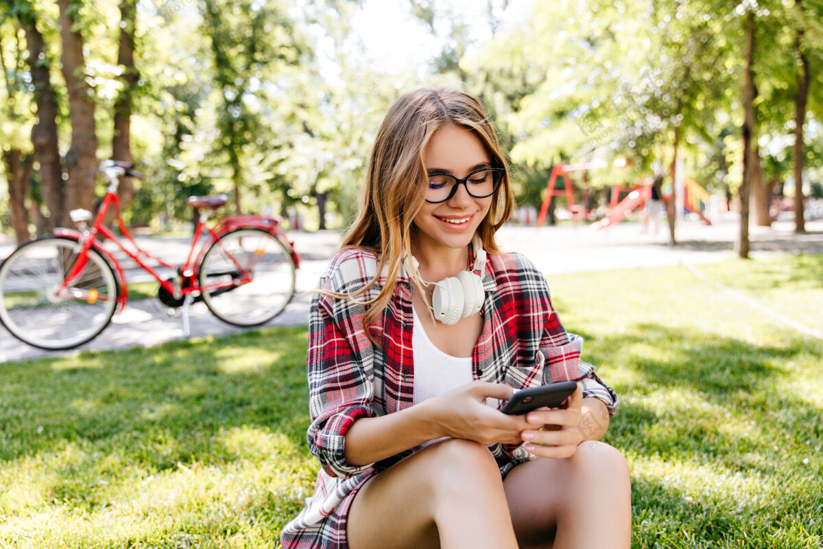 快乐幸福的白人女士在公园里打电话戴着休闲眼镜的快乐的欧洲女孩坐在草地上发短信女性人乡村