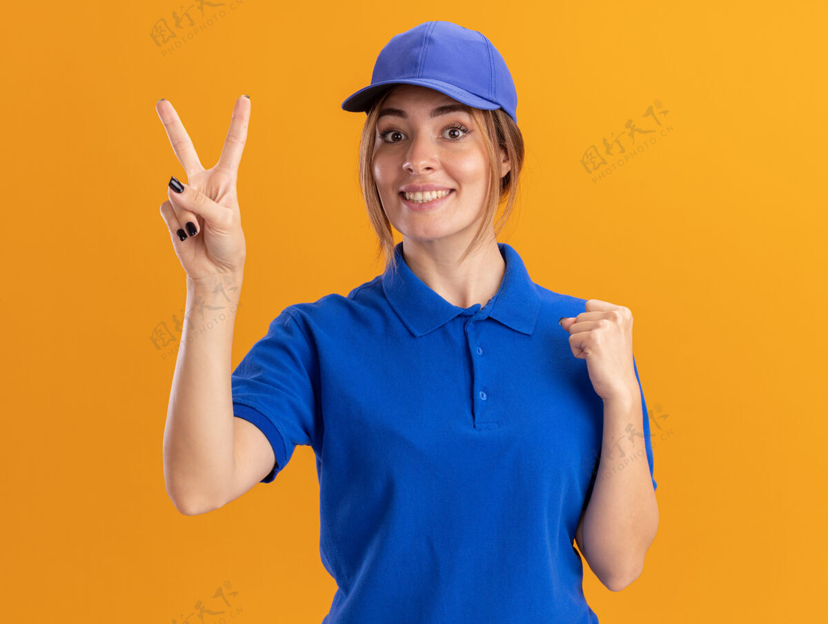 漂亮微笑着的年轻漂亮的女送货员穿着制服保持拳头和手势胜利的手上橙色的标志拳头胜利传递