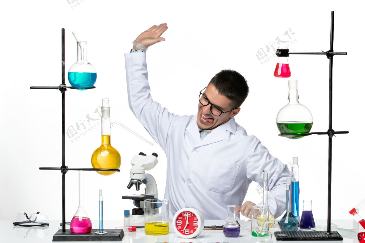 男性前视图穿着白色医疗服的男性化学家坐在浅白背景的病毒科学冠状病毒大流行实验室里拿着溶液解决方案实验室人