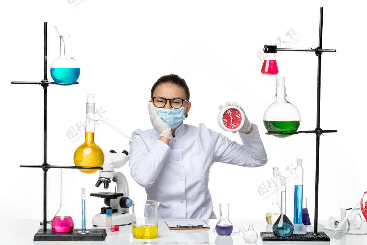 保持正面图白色办公桌上戴着面具的女化学家拿着时钟病毒实验室化学冠状病毒-飞溅面具喷溅外套