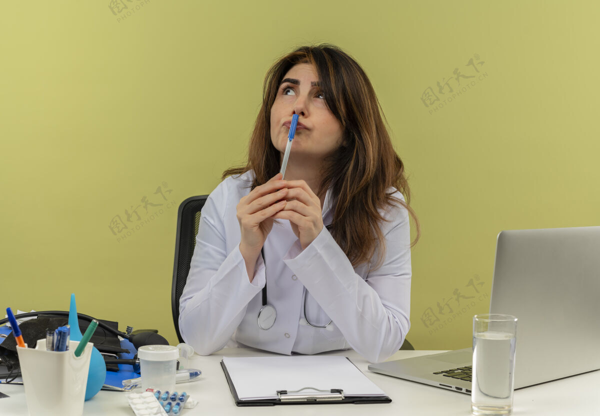 起来体贴的中年女医生穿着医用长袍和听诊器坐在办公桌旁 拿着医疗工具 剪贴板和笔记本电脑 抬起头来 用笔触摸嘴唇听诊器成人女性