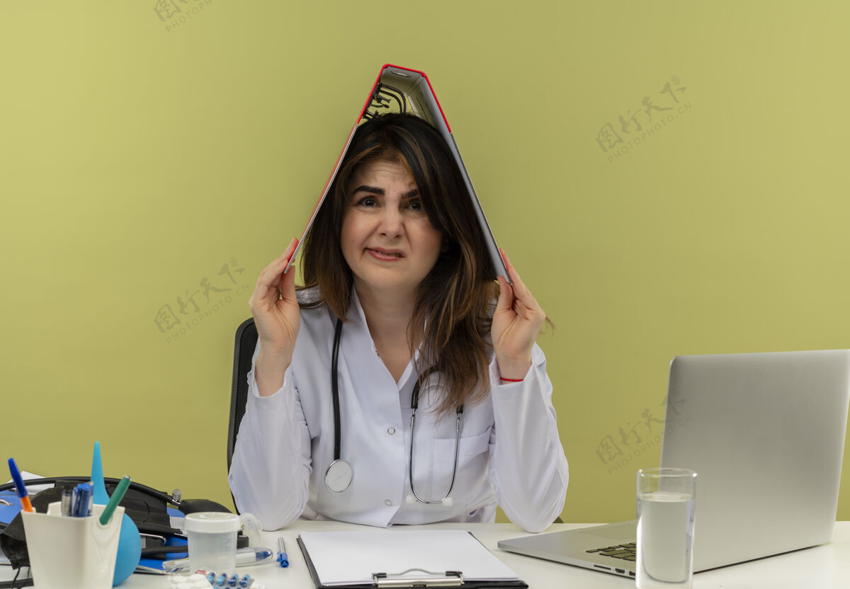女士未出租的中年女医生穿着医用长袍和听诊器坐在办公桌旁 头上拿着医疗工具和笔记本电脑夹人文件夹成人