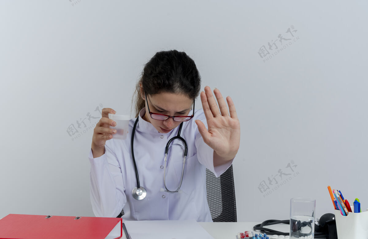 成人年轻的女医生穿着医用长袍 戴着听诊器 戴着眼镜 坐在办公桌旁 拿着医疗工具 拿着医用烧杯 低头做着停止手势烧杯女士听诊器