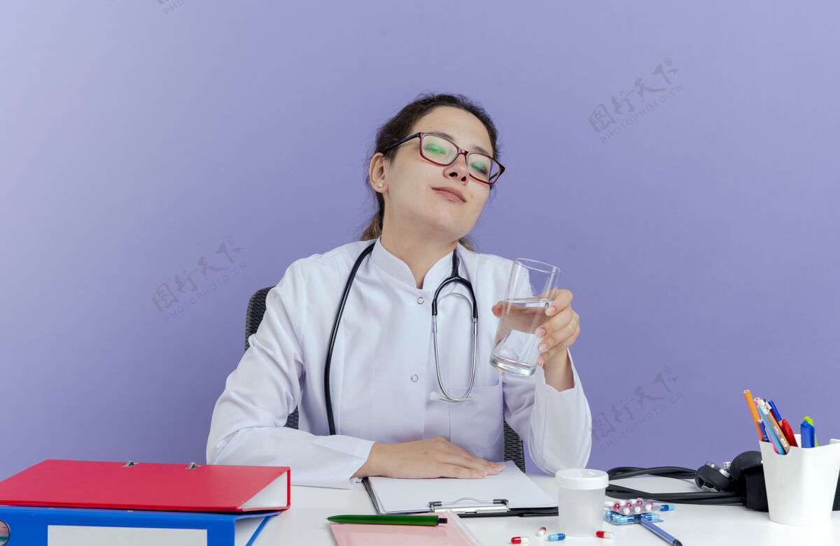 医疗高兴的年轻女医生穿着医用长袍和听诊器坐在办公桌旁 手里拿着医疗工具望着一杯水桌子穿紫色