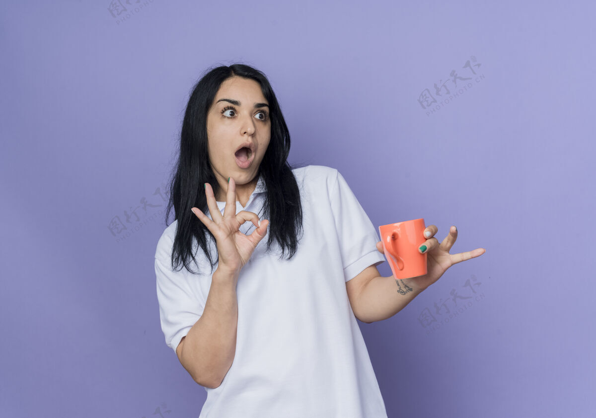 手自信的年轻黑发白种女孩拿着杯子 摆出ok手势 看着紫色墙上孤立的一面黑发好的自信