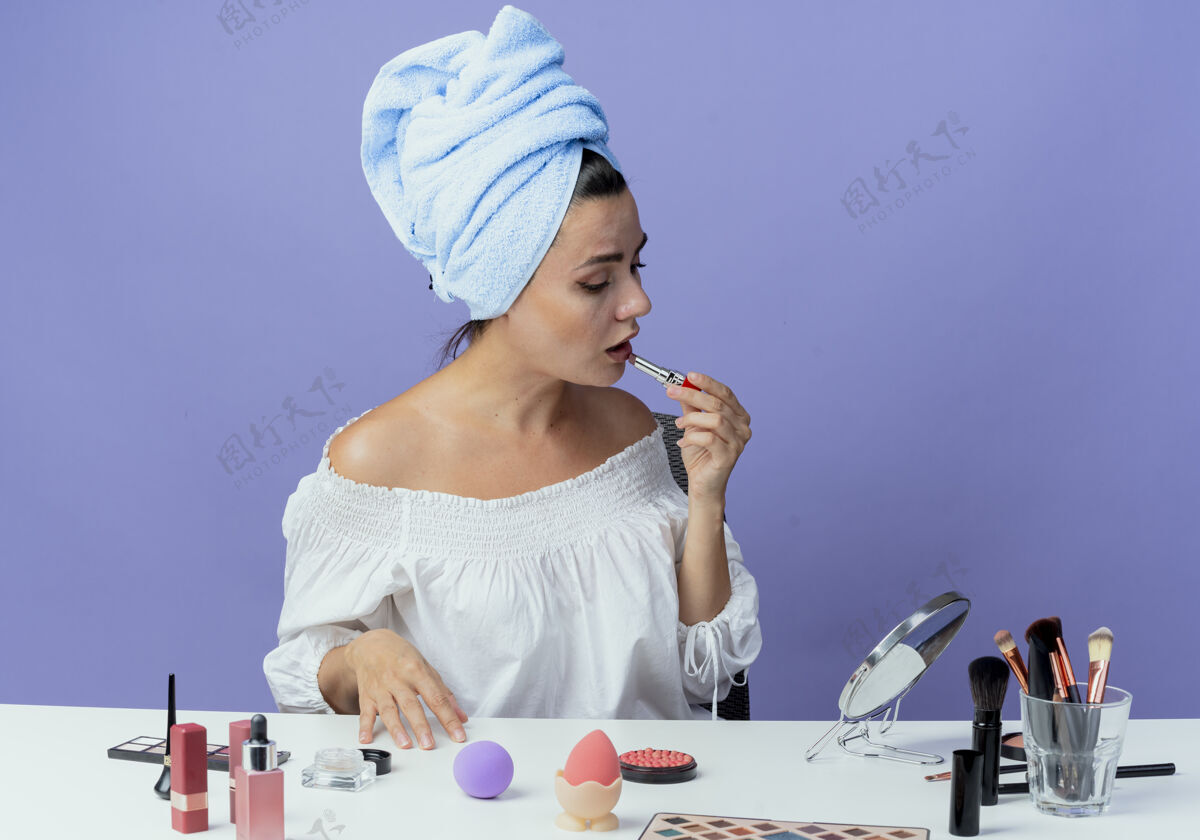 桌子美丽的心烦意乱的女孩裹着发巾坐在桌子上 拿着化妆工具 拿着唇膏 看着镜子在紫墙上孤立不舒服坐毛巾