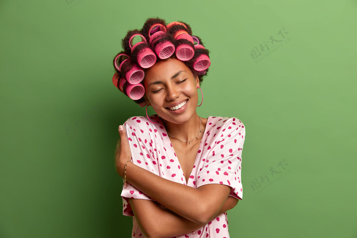 拥抱高兴的黑皮肤女模特拥抱自己 喜欢穿柔软的睡衣 仰着头 愉快地微笑 戴着做完美发型的发夹 孤立在绿色上心情女人舒适
