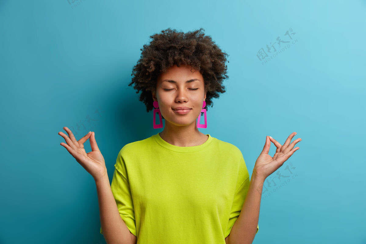姿势冷静放松的黑皮肤女人站在莲花的姿势 感到轻松 试图集中在和平的气氛中 穿着休闲的绿色衣服 隔离在蓝色的墙上肢体语言的概念成人愉悦情绪